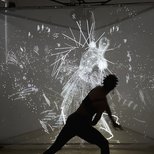 A silhouetted figure moves in front of a glowing projection of abstract, motion-reactive visuals forming a ghostly human shape.