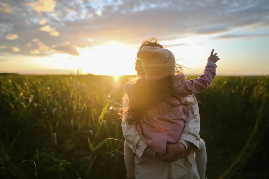 Person holding a child in a field at sunset, with the sun casting a warm glow over tall grass.