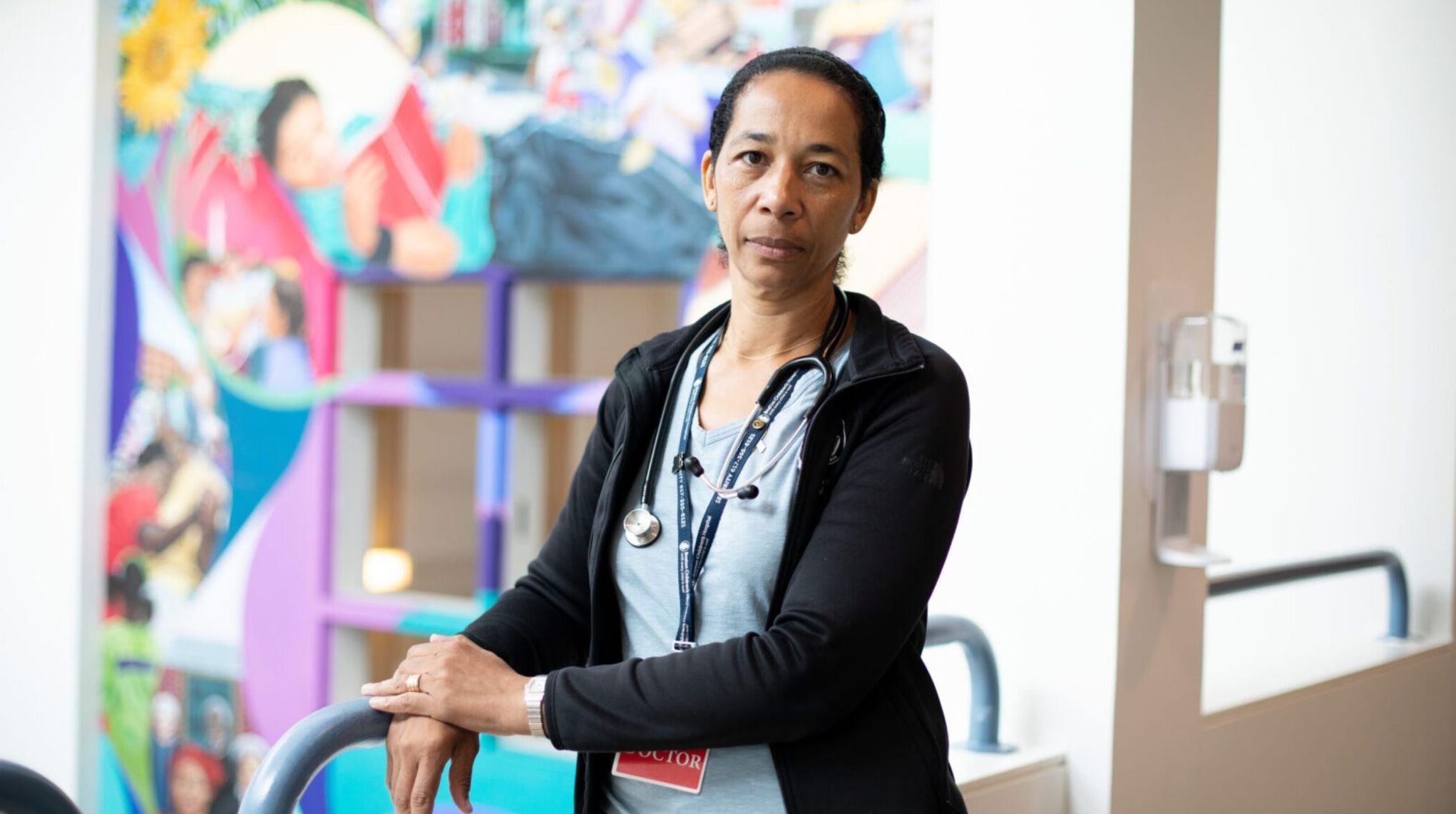 Healthcare provider with a stethoscope stands in a hospital hallway in front of a colorful mural.