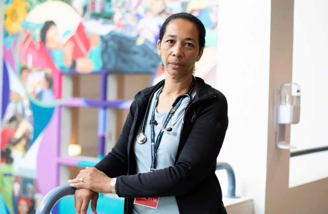 Healthcare provider with a stethoscope stands in a hospital hallway in front of a colorful mural.