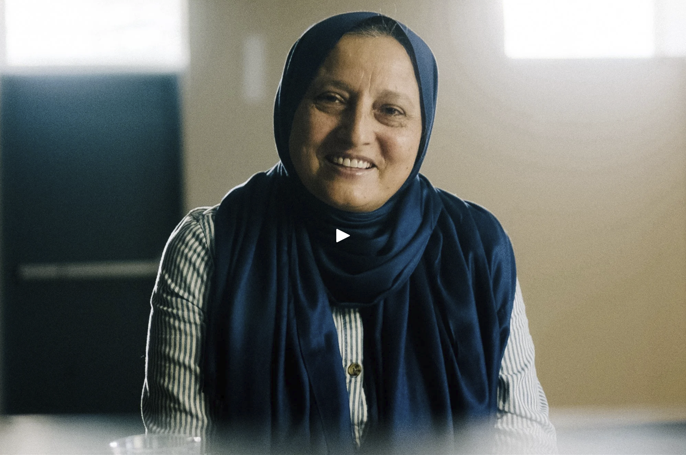 Smiling woman in a headscarf appears in a paused video frame, seated in an interview-style setting with soft lighting.