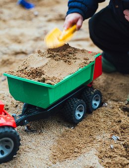 Close-up of a green toy dump truck filled with sand; a small hand is holding a yellow shovel