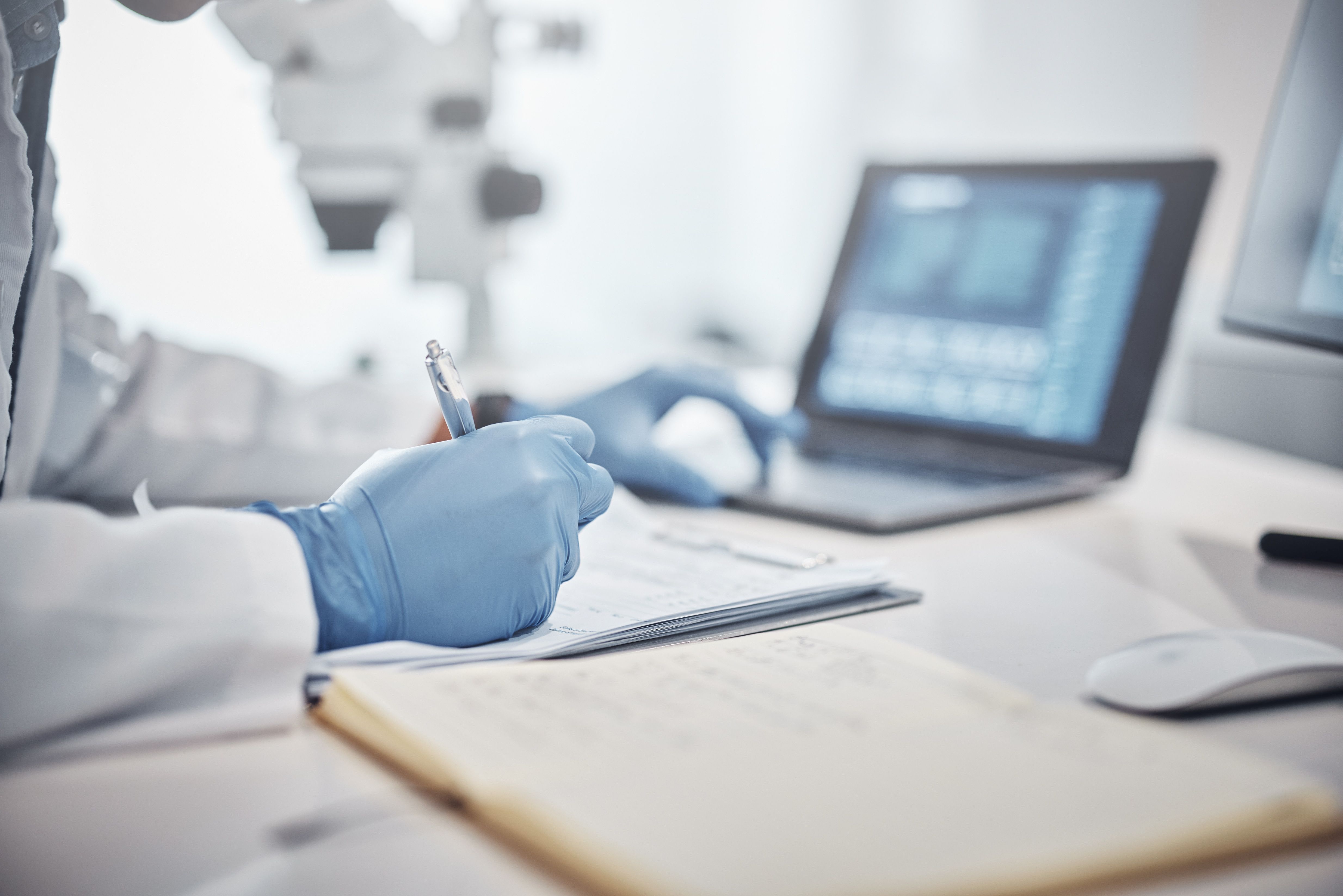 Medical researcher in a white lab coat and blue gloves writes notes in a laboratory with microscopes and a laptop in the background.