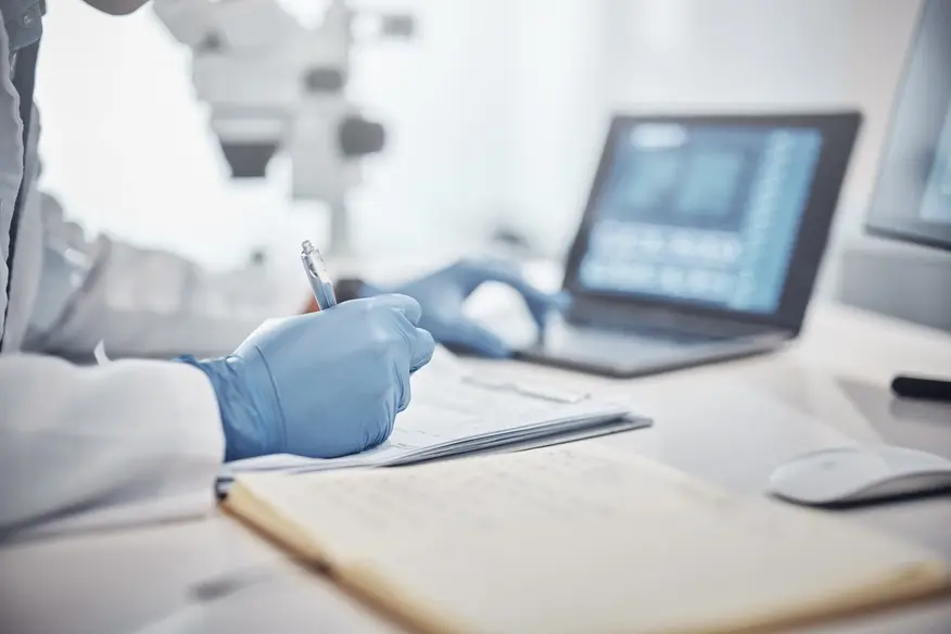 Medical researcher in a white lab coat and blue gloves writes notes in a laboratory with microscopes and a laptop in the background.