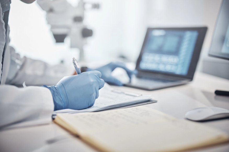 Medical researcher in a white lab coat and blue gloves writes notes in a laboratory with microscopes and a laptop in the background.