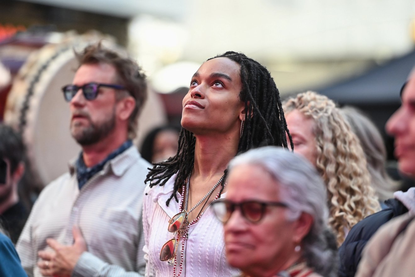 Woman in a striped shirt looks upward in a crowd during an outdoor event, surrounded by people and a drum in the background.