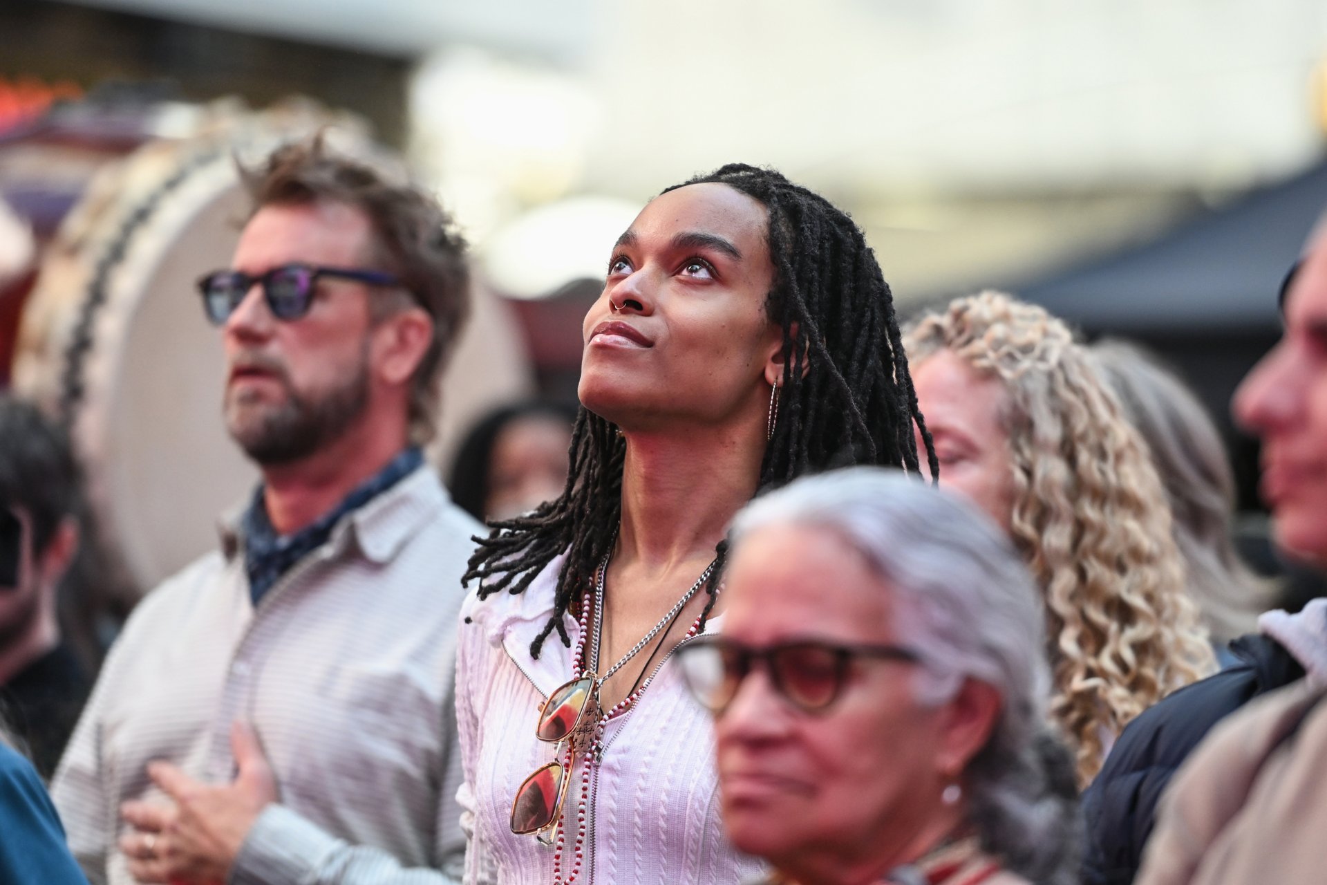 Woman in a striped shirt looks upward in a crowd during an outdoor event, surrounded by people and a drum in the background.