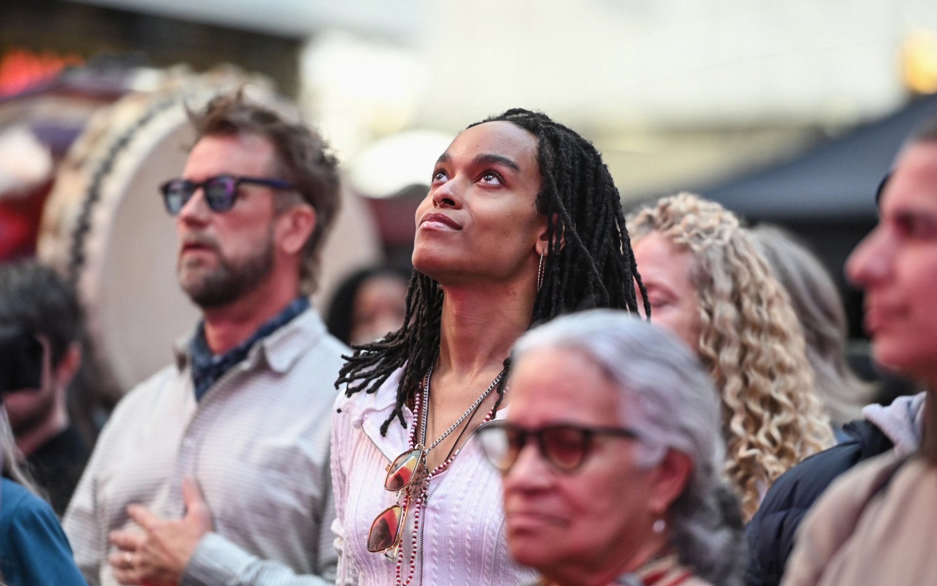 Woman in a striped shirt looks upward in a crowd during an outdoor event, surrounded by people and a drum in the background.