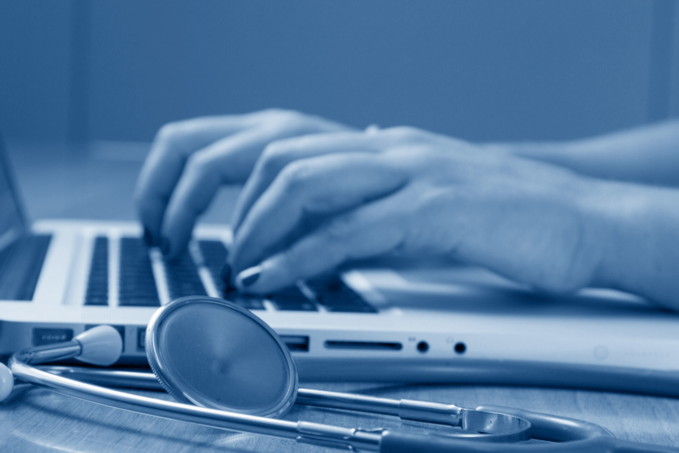 Person typing on a laptop with a stethoscope nearby, in a blue monochrome color scheme.