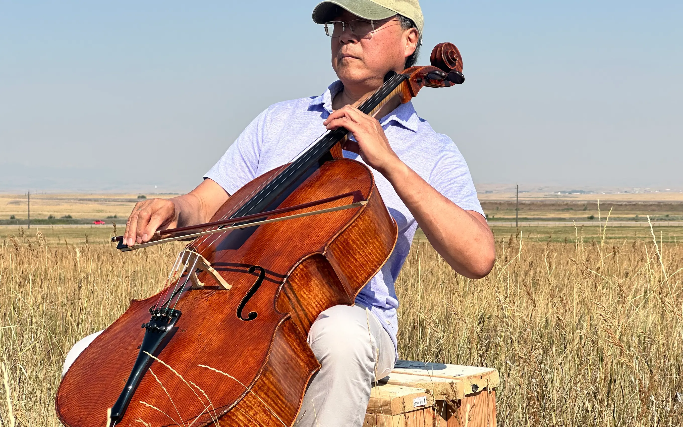 Musician plays a cello while seated in a grassy prairie landscape.