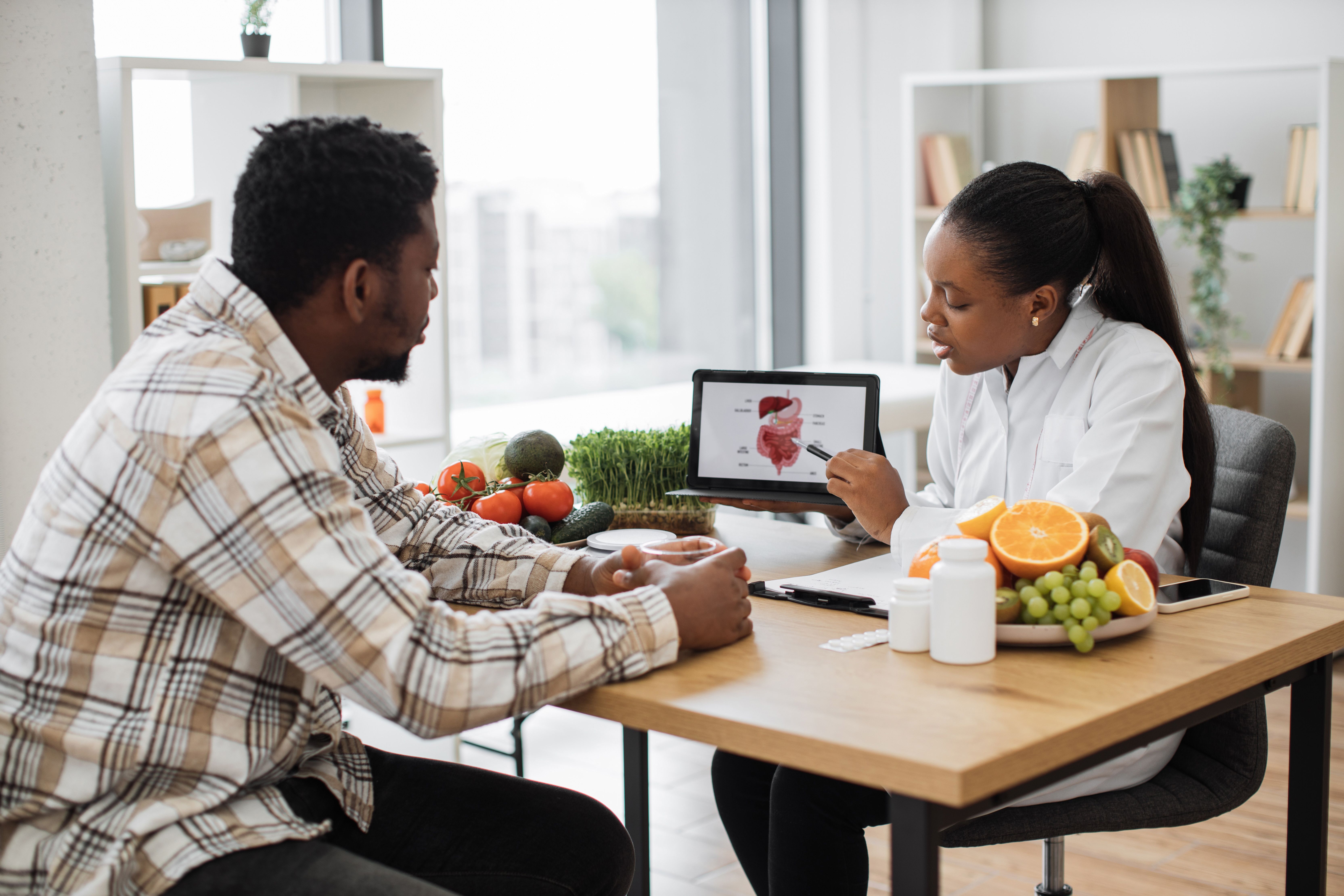 Nutritionist consulting with a patient at a table with fresh fruits, vegetables, and a tablet showing health information.