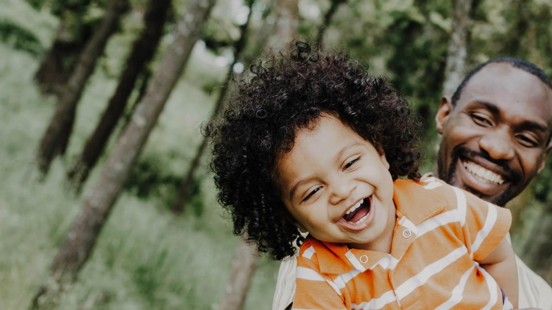 Smiling father holds his laughing young child while outdoors in a green, tree-lined park.