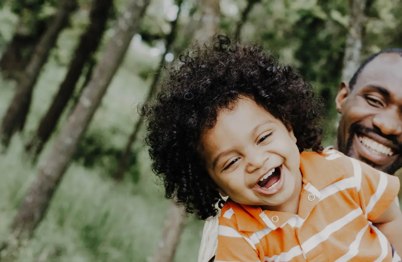 Smiling father holds his laughing young child while outdoors in a green, tree-lined park.
