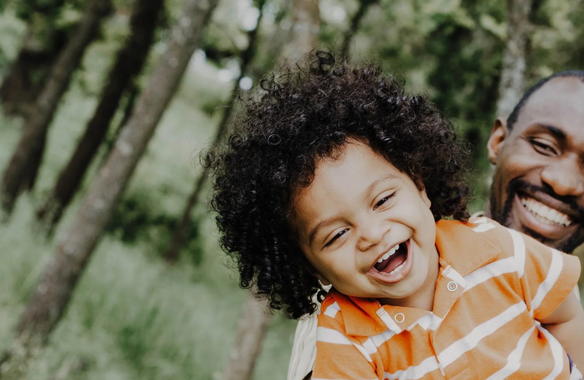 Smiling father holds his laughing young child while outdoors in a green, tree-lined park.