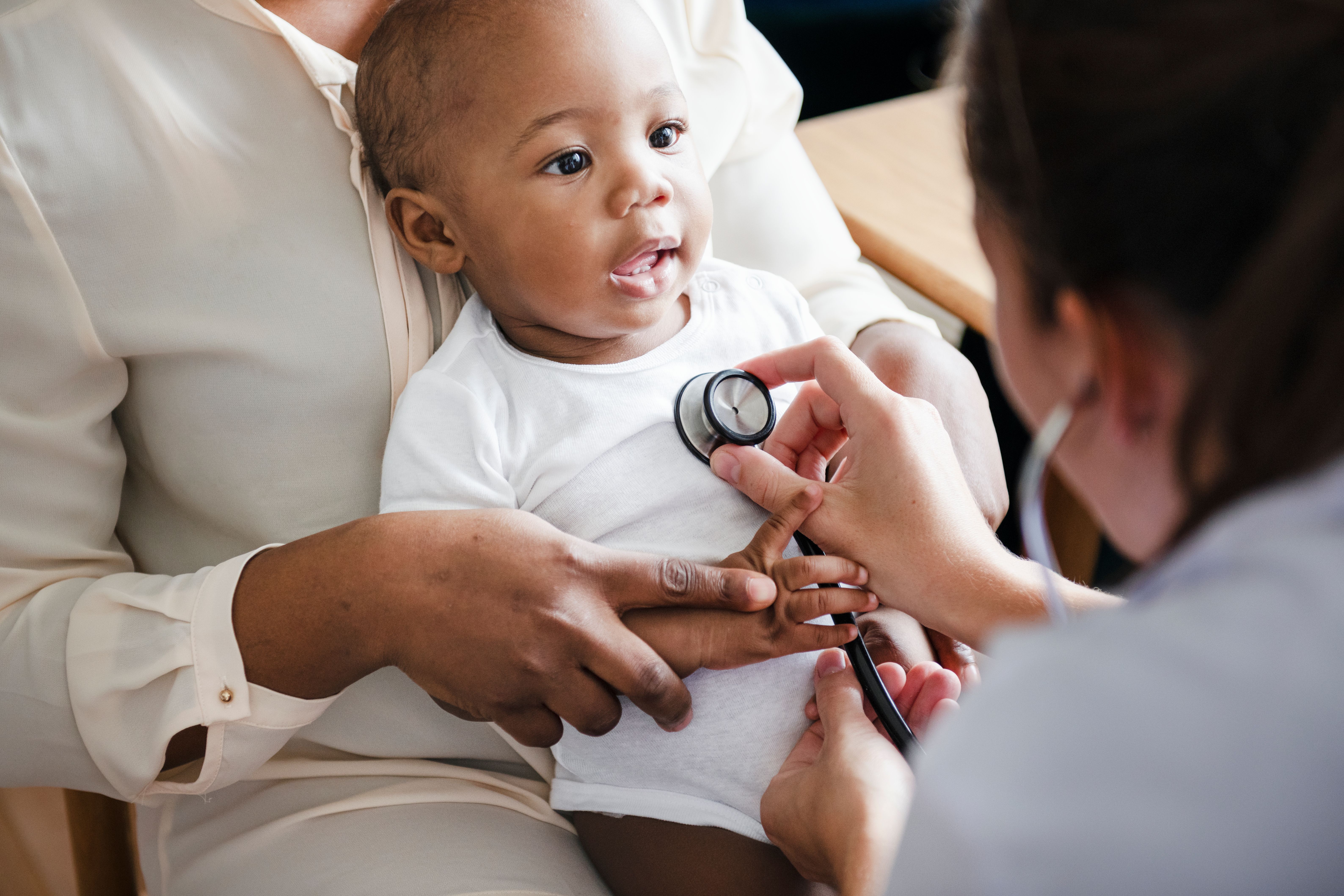 Smiling Black infant in a white onesie sitting on a caregiver’s lap during a pediatric visit.
