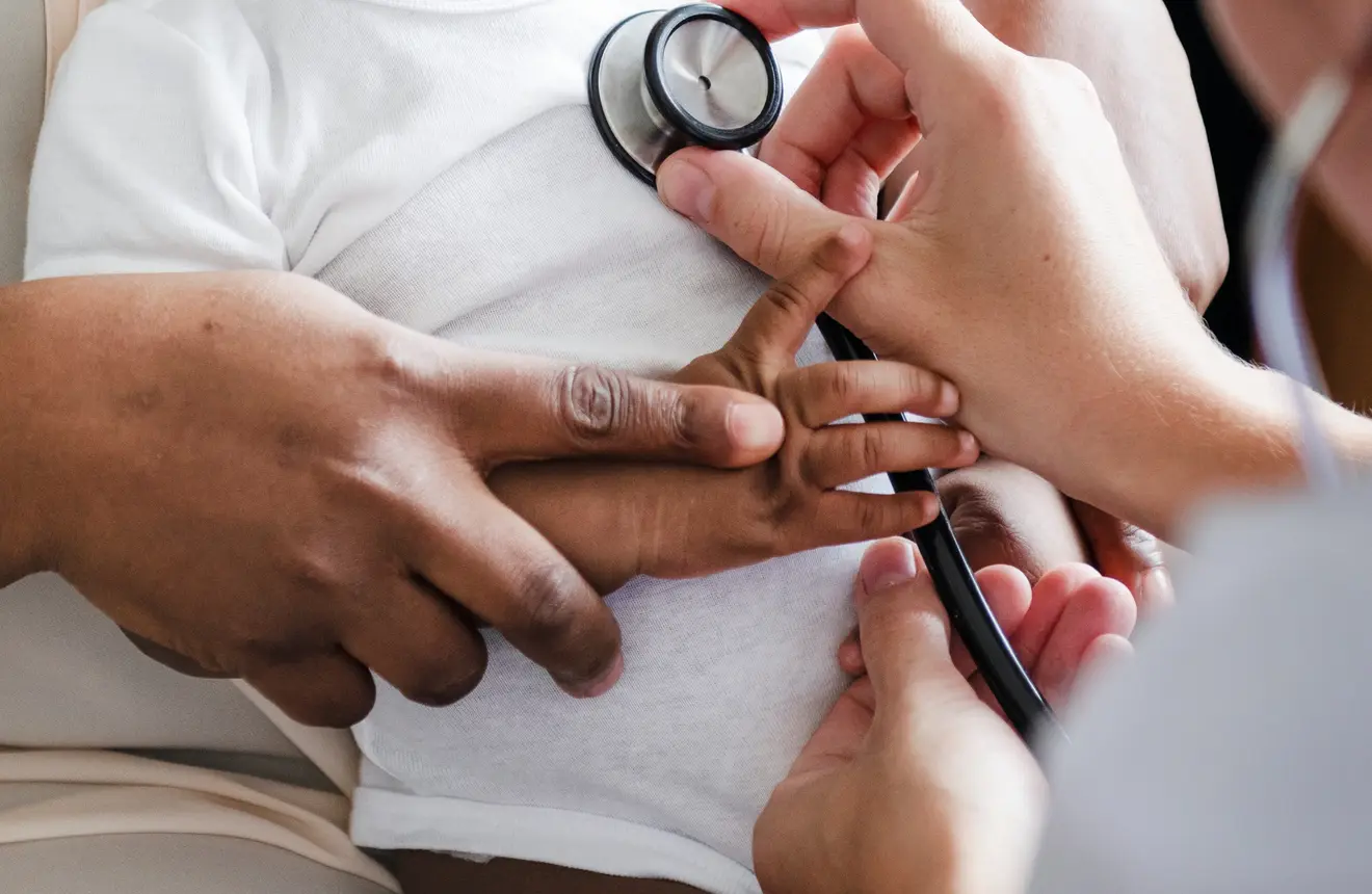 Smiling Black infant in a white onesie sitting on a caregiver’s lap during a pediatric visit.