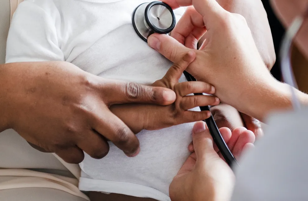 Smiling Black infant in a white onesie sitting on a caregiver’s lap during a pediatric visit.