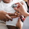 Smiling Black infant in a white onesie sitting on a caregiver’s lap during a pediatric visit.