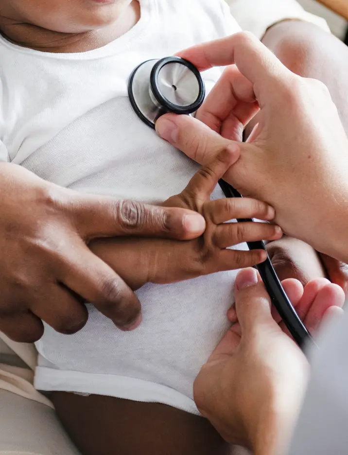 Smiling Black infant in a white onesie sitting on a caregiver’s lap during a pediatric visit.