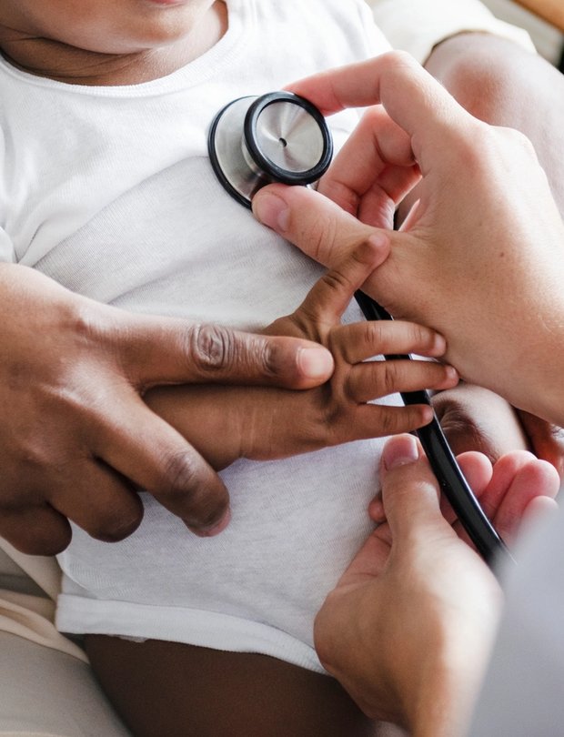 Smiling Black infant in a white onesie sitting on a caregiver’s lap during a pediatric visit.