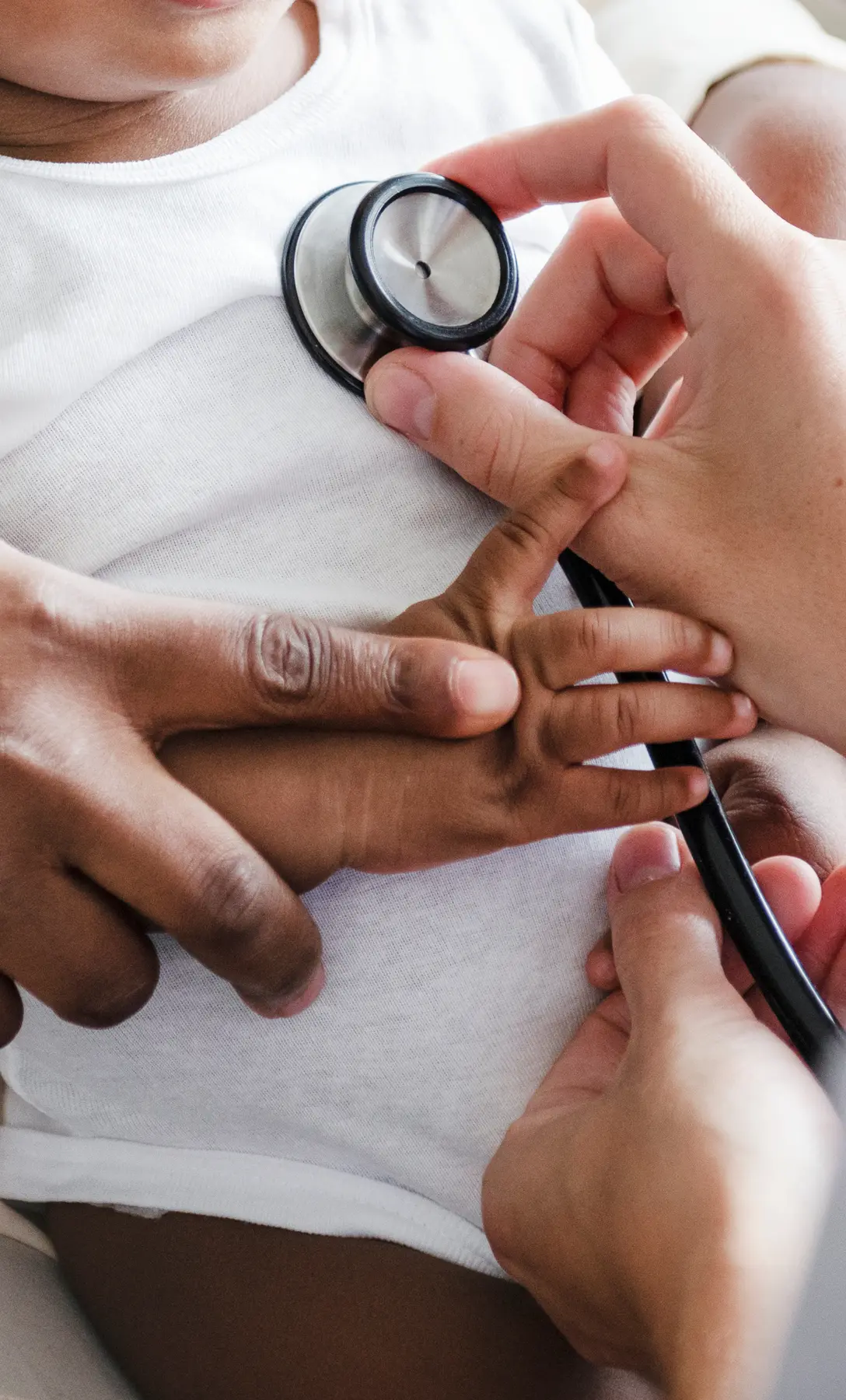 Smiling Black infant in a white onesie sitting on a caregiver’s lap during a pediatric visit.