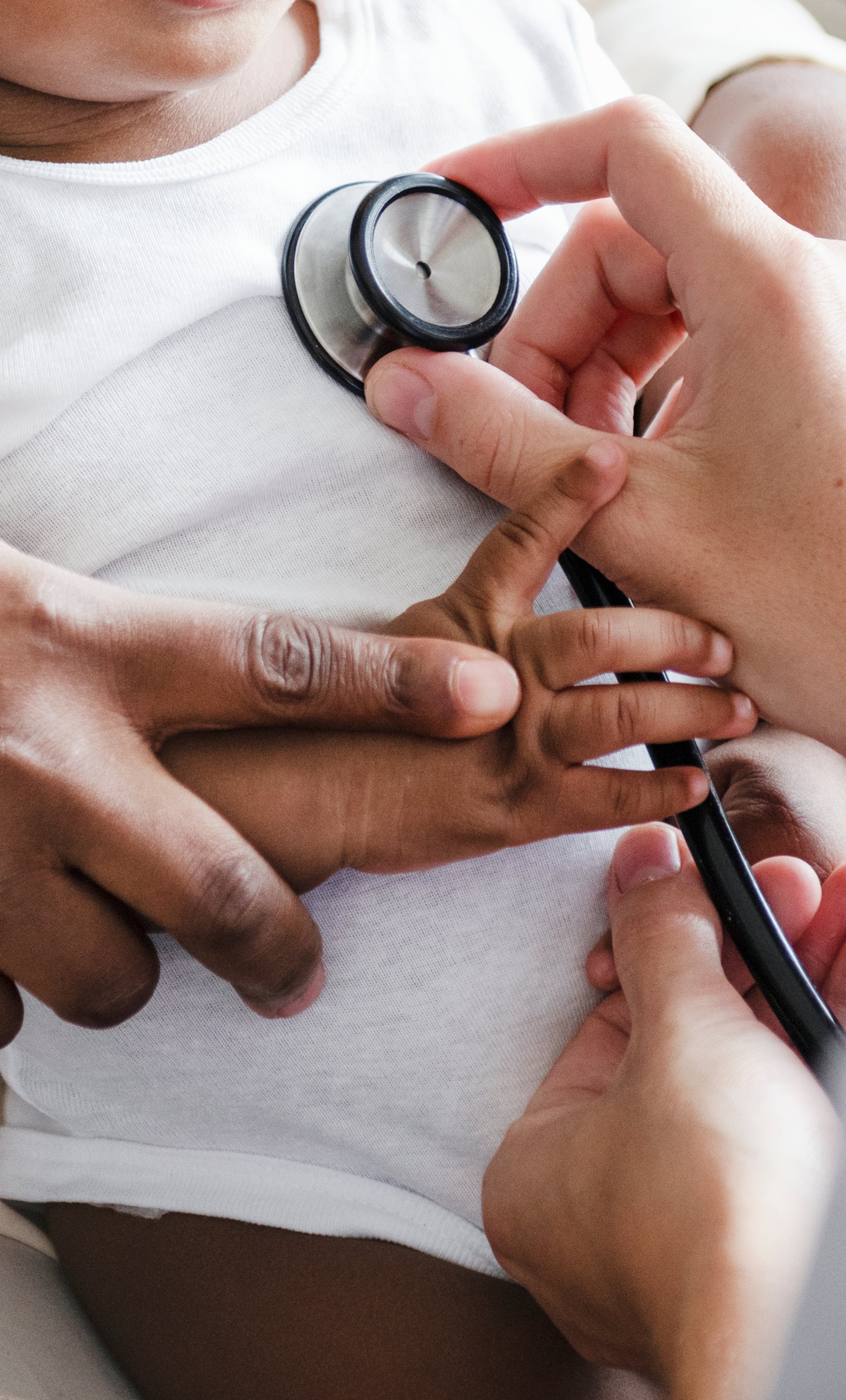 Smiling Black infant in a white onesie sitting on a caregiver’s lap during a pediatric visit.