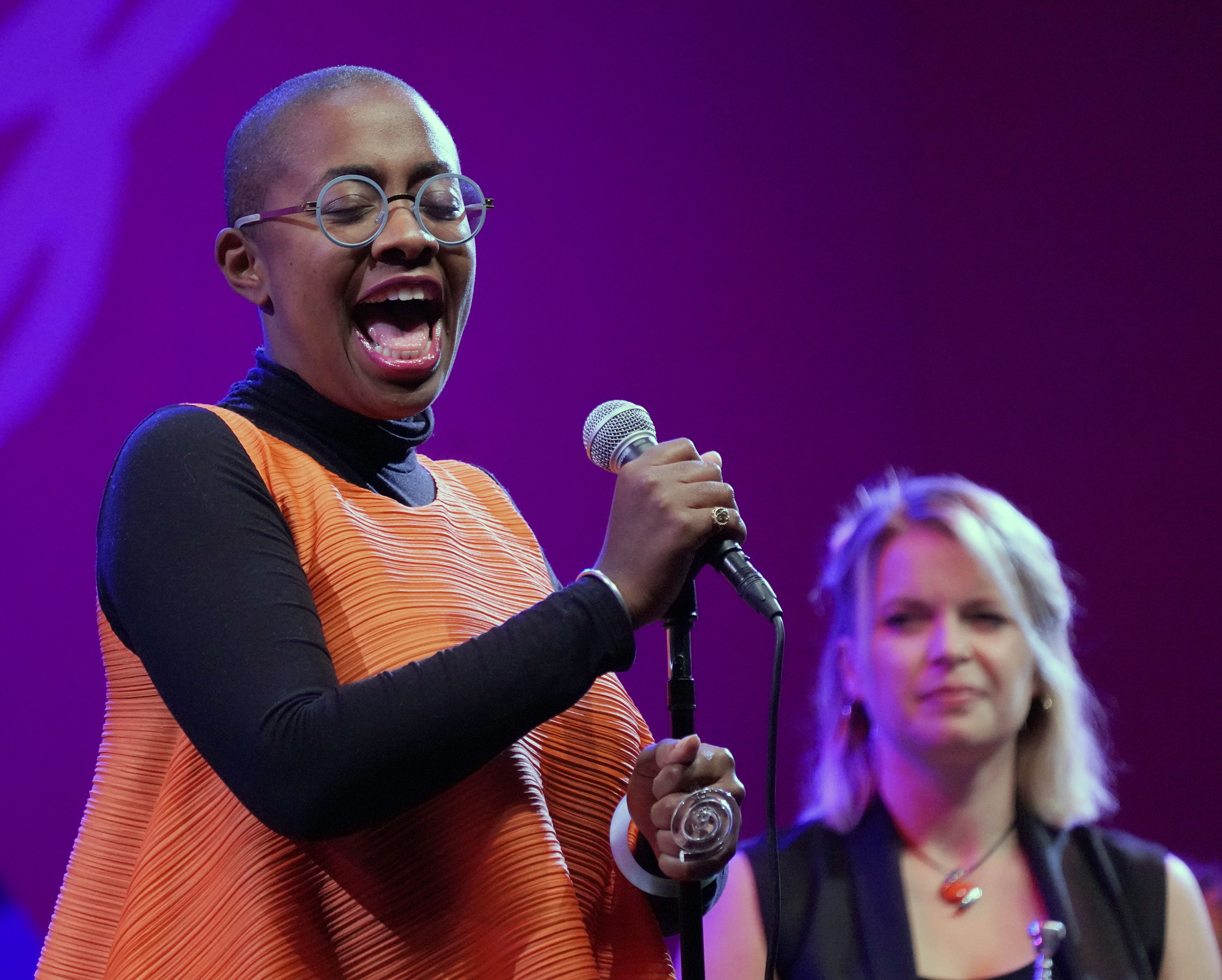 Jazz vocalist Cécile McLorin Salvant sings into a microphone onstage, with a purple-lit background and a backup singer behind her.