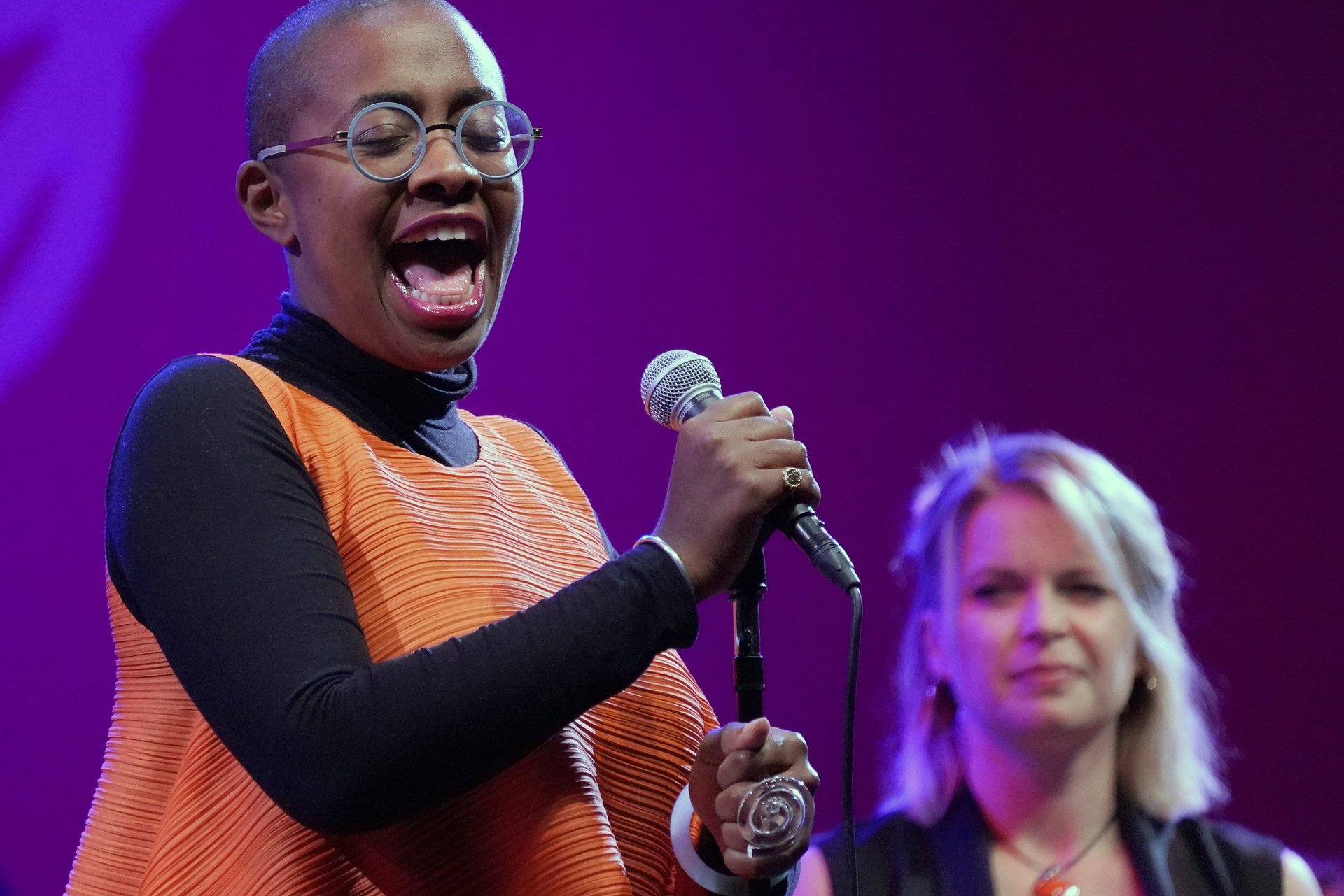 Jazz vocalist Cécile McLorin Salvant sings into a microphone onstage, with a purple-lit background and a backup singer behind her.