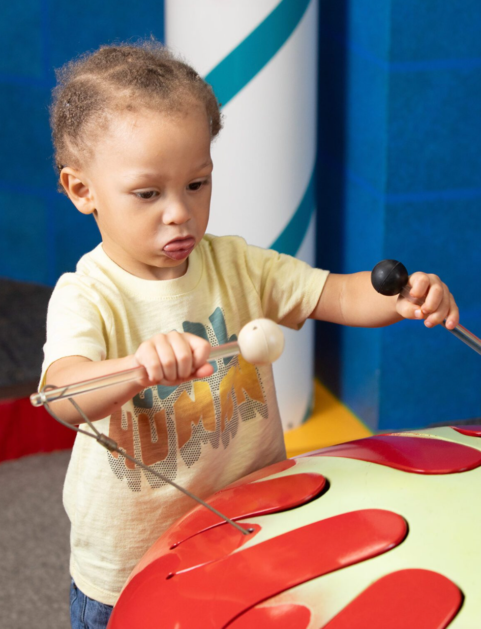 Young child uses two mallets to play a colorful drum in an indoor play area.