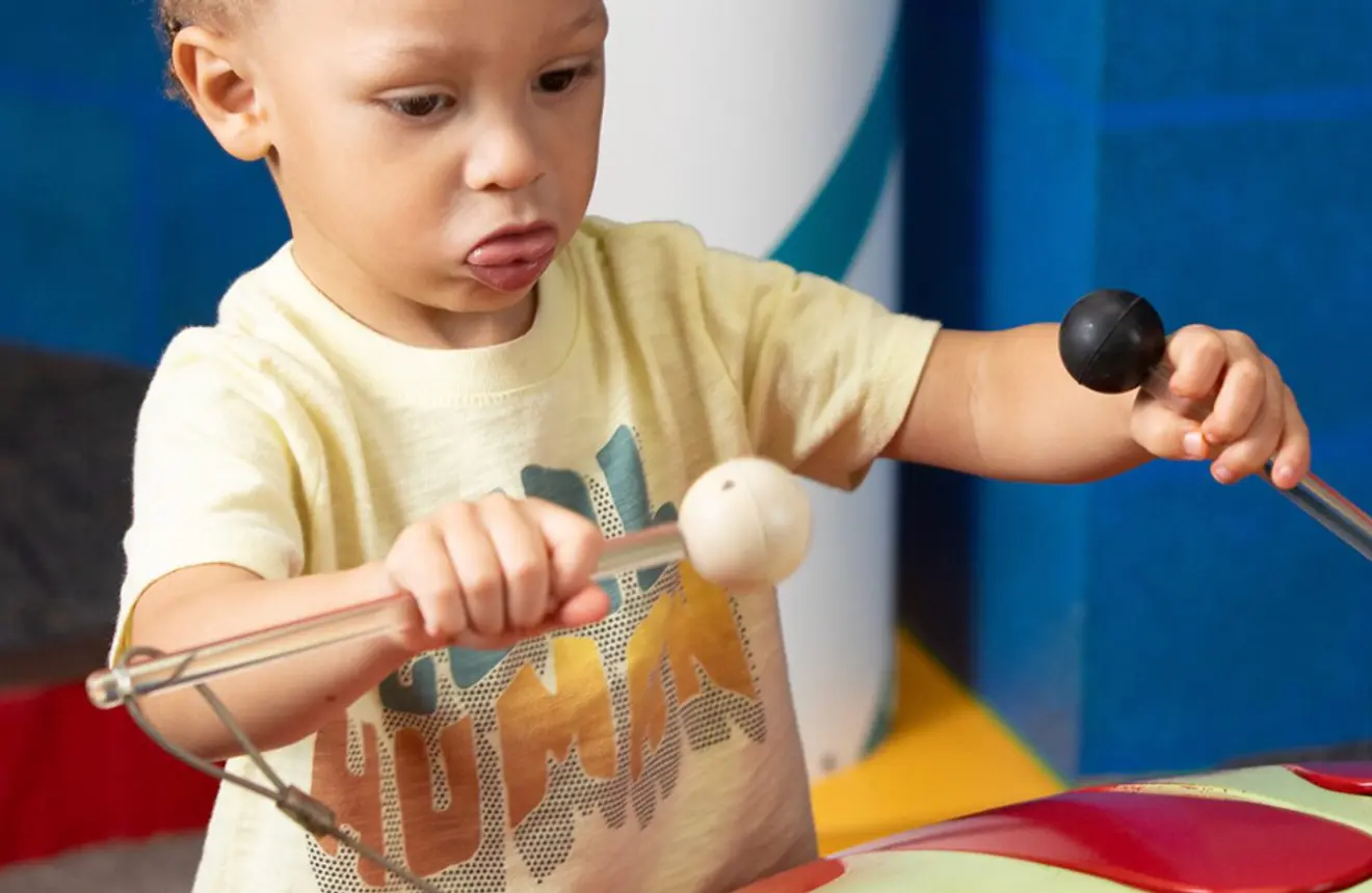 Young child uses two mallets to play a colorful drum in an indoor play area.