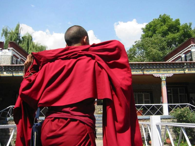 A Tibetan Buddhist monk dressed in traditional maroon and red robes walking toward a stone building with mountain architecture in McLeod Ganj, Dharamshala.
