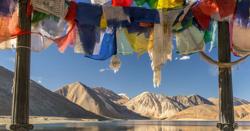 Prayer flags at Pangong Tso shoreline with tent camps visible — overnight camp accommodation along the Indian shoreline costs ₹2,000–₹5,000 per person including dinner and breakfast; power cuts at 10pm; the pre-dawn stillness makes it worth every rupee