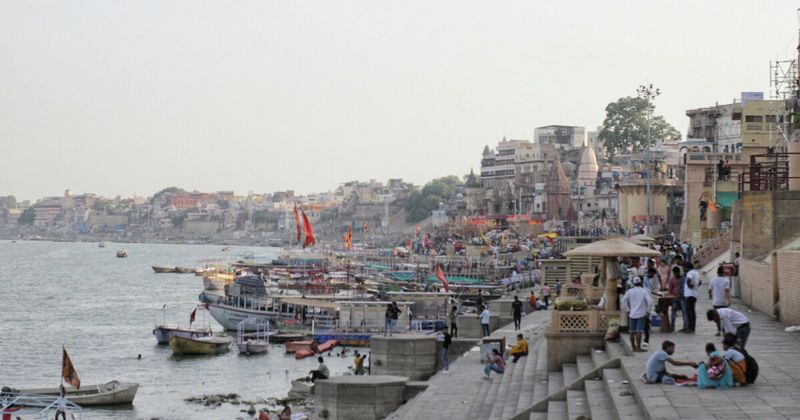 A wooden rowboat on the Ganges with the Varanasi ghats visible across the full background — the perspective from the river is how most first-time visitors understand the city's scale and continuity for the first time