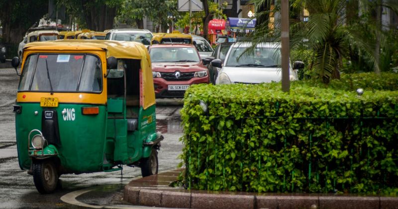 A vibrant yellow and green three-wheeled auto-rickshaw parked on a busy, sunlit street in India, featuring its open-sided cabin and black vinyl seats.