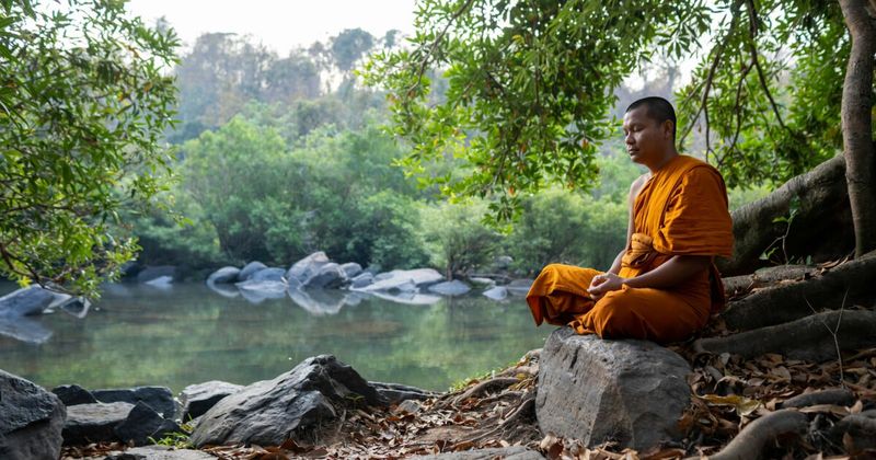 A Tibetan Buddhist monk in traditional maroon robes sitting in meditation on a large river rock by a flowing mountain stream in Dharamshala, Himachal Pradesh.