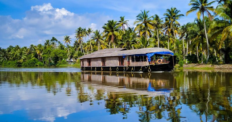 Traditional Kerala houseboat gliding through narrow Alleppey backwater canals lined with coconut palms — overnight deluxe houseboats start from ₹8,500 for two persons including all meals and crew
