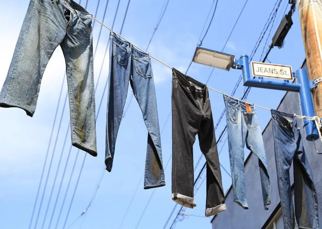 Multiple pairs of jeans hanging on a clothesline beneath a "JEANS St." street sign, creating a visual pun as denim meets its namesake street.