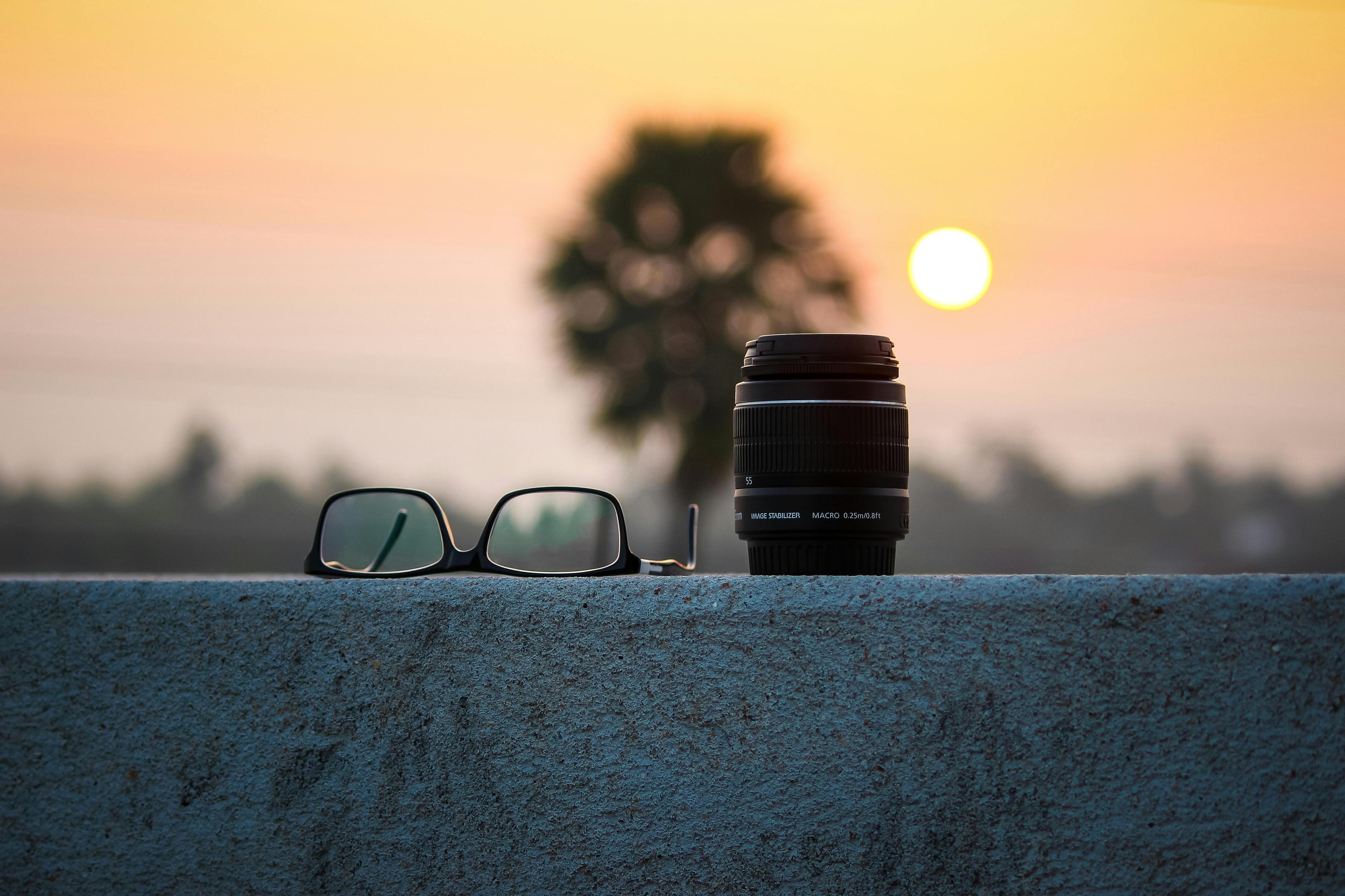 Kameraobjektiv und Brille auf einer Mauer vor einem Sonnenuntergang.