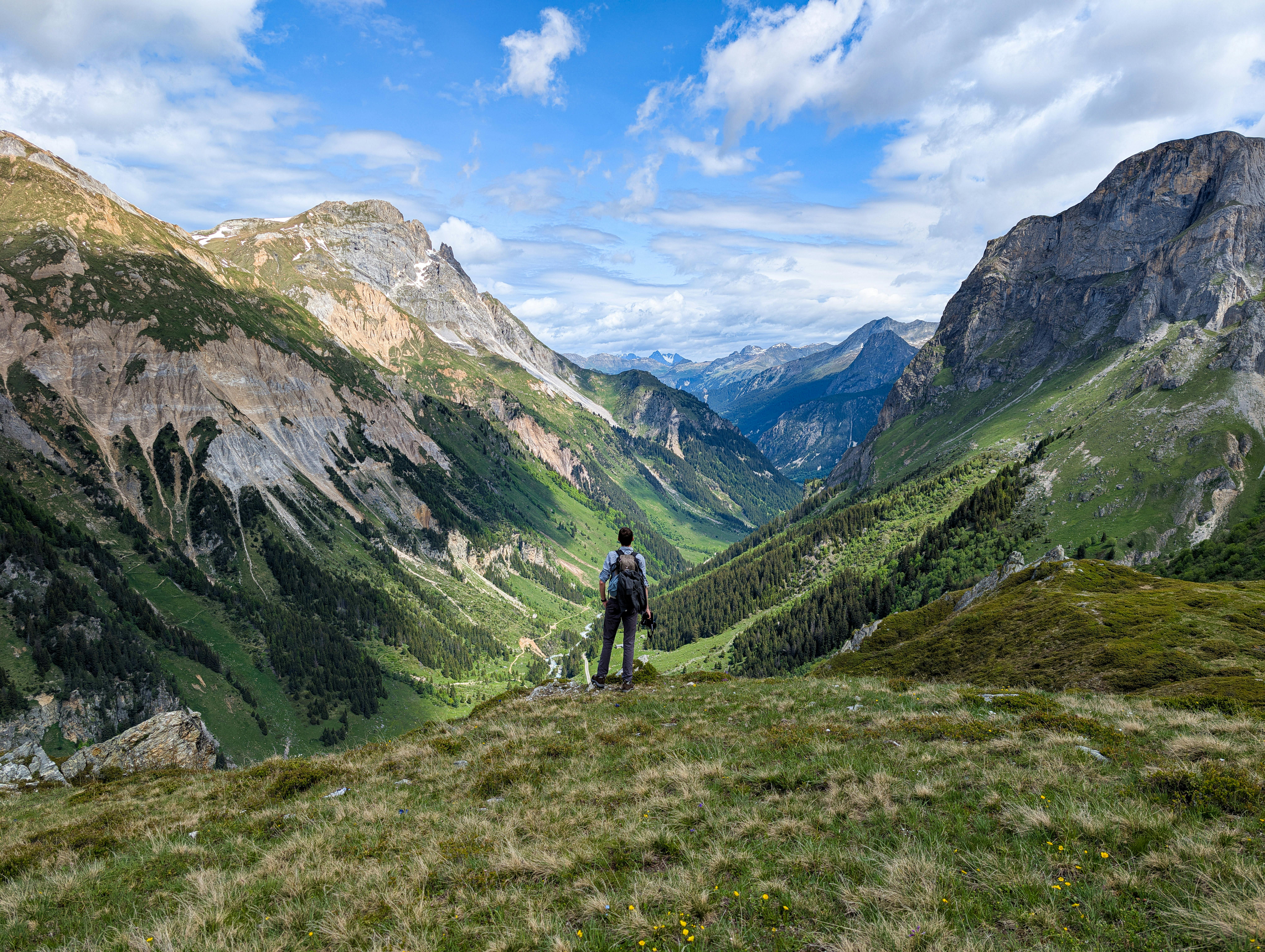 Person steht auf einer Wiese mit Rucksack und blickt auf hohe grüne Berge.