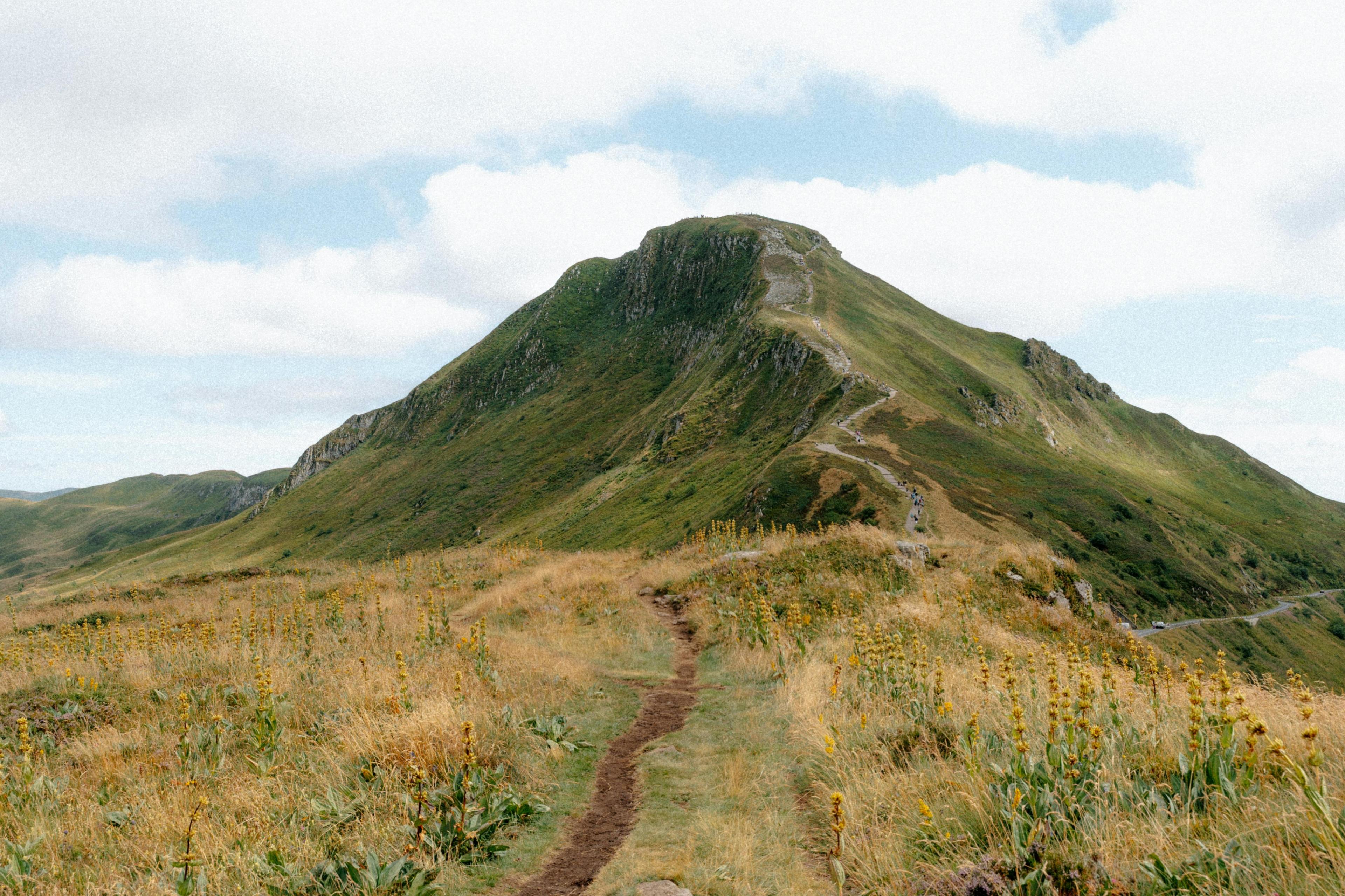 Schmaler Wanderpfad verläuft durch eine grasbewachsene Berglandschaft hinauf zu einem markanten Gipfel unter leicht bewölktem Himmel.