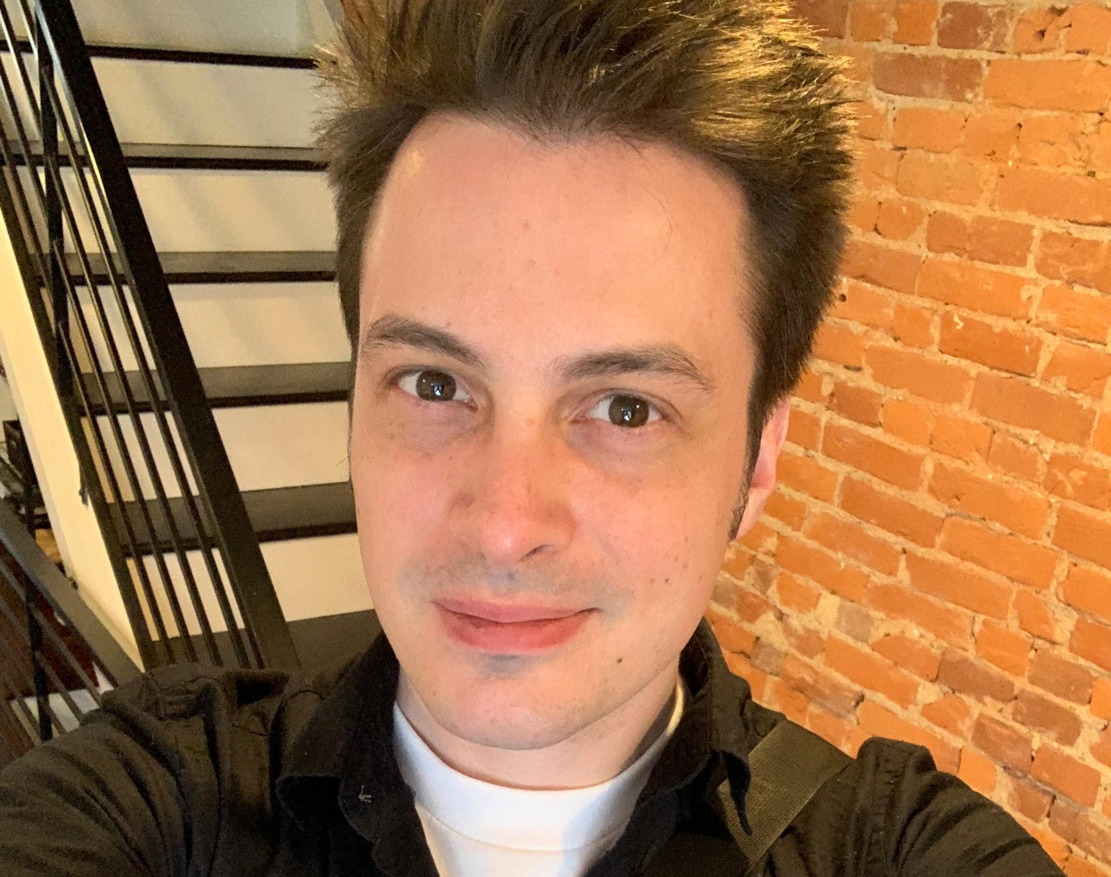 Andrew Cunningham, writer at Tailscale, wearing a black shirt on a white undershirt, against a black and white stairway and brick wall.