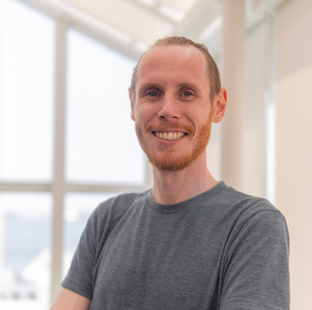 Man with short beard wearing a grey t-shirt in front of brightly lit windows.