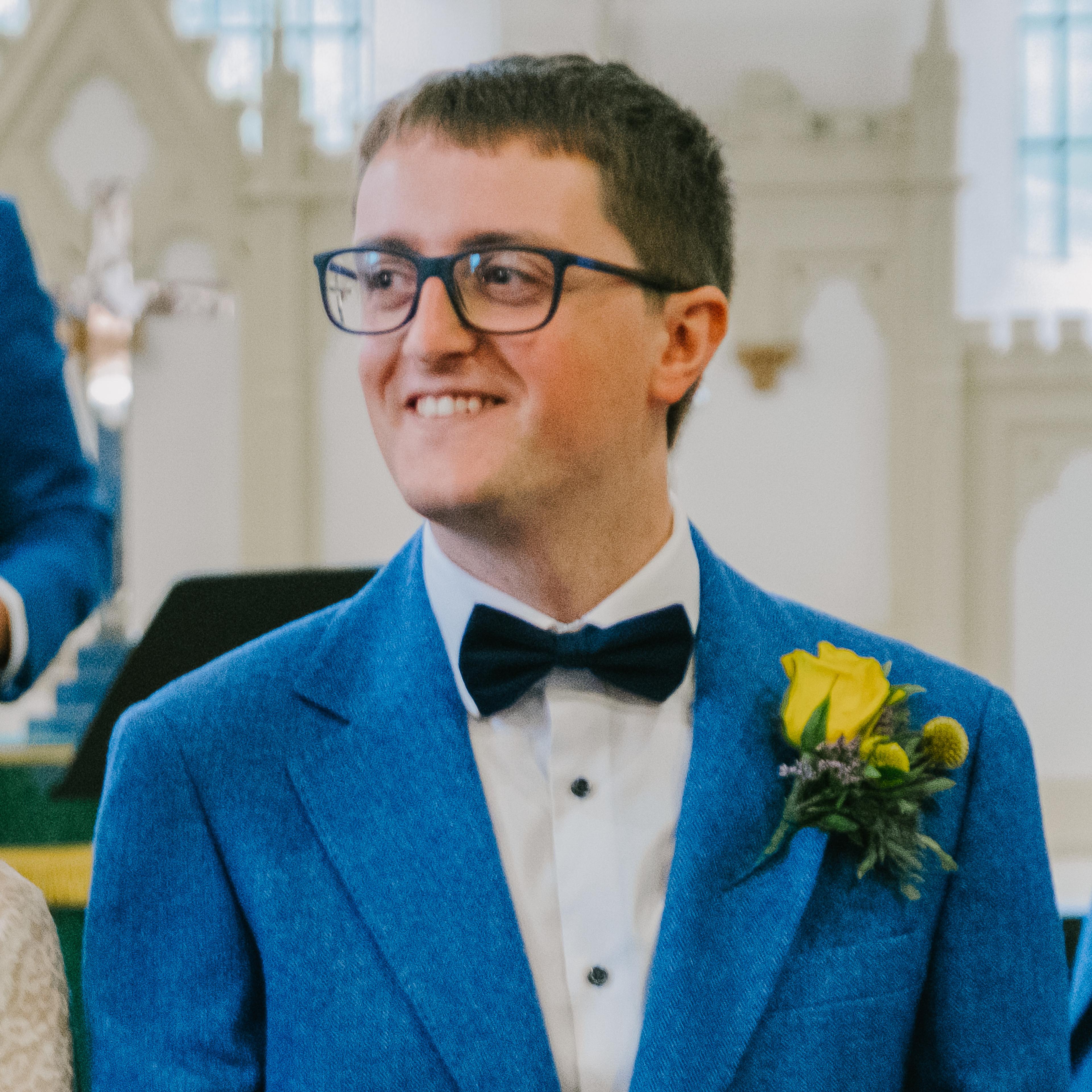 A smiling man wearing glasses, a bow tie, and a boutonnière with a rose, dressed in a formal suit, standing in a light-filled room.