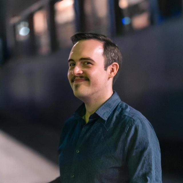 A person with short dark hair, wearing a dark button-up shirt, smiling slightly while standing in front of a blurred urban background at night.