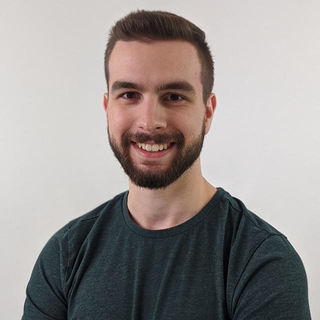 A man smiling and facing the camera. He has short, neatly trimmed hair and a full beard.