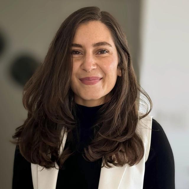 A woman with long, wavy brown hair and a warm smile is looking directly at the camera. She is wearing a black turtleneck sweater and a white vest. The background is softly blurred with neutral tones.
