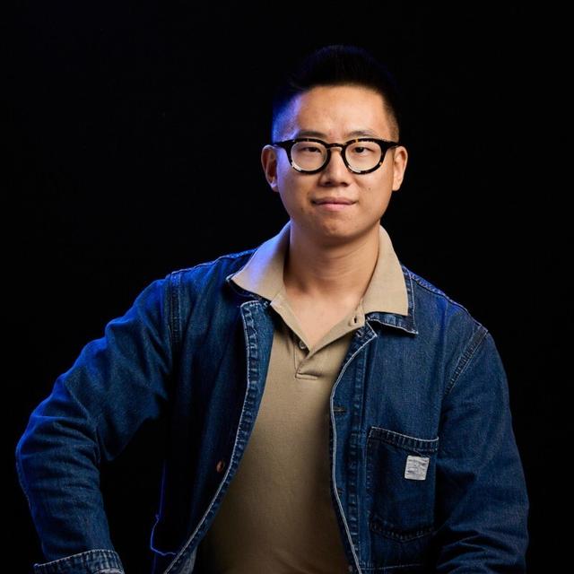 A man wearing glasses, a tan collared shirt, and a denim jacket sits against a dark background, looking at the camera with a slight smile.