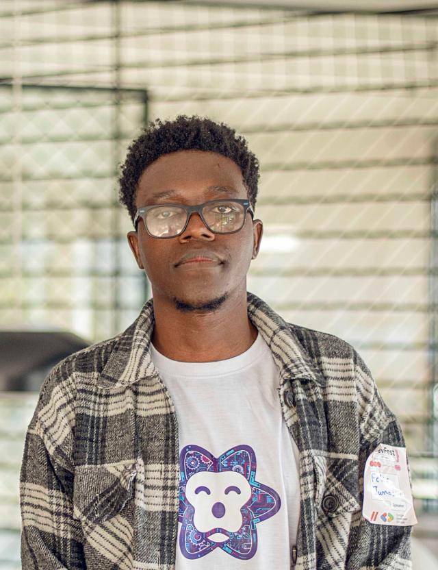 A person wearing glasses stands facing the camera indoors. They have short, curly hair and are wearing a white t-shirt with a design under a plaid shirt. The background is softly blurred with metal railings and a light, industrial interior.