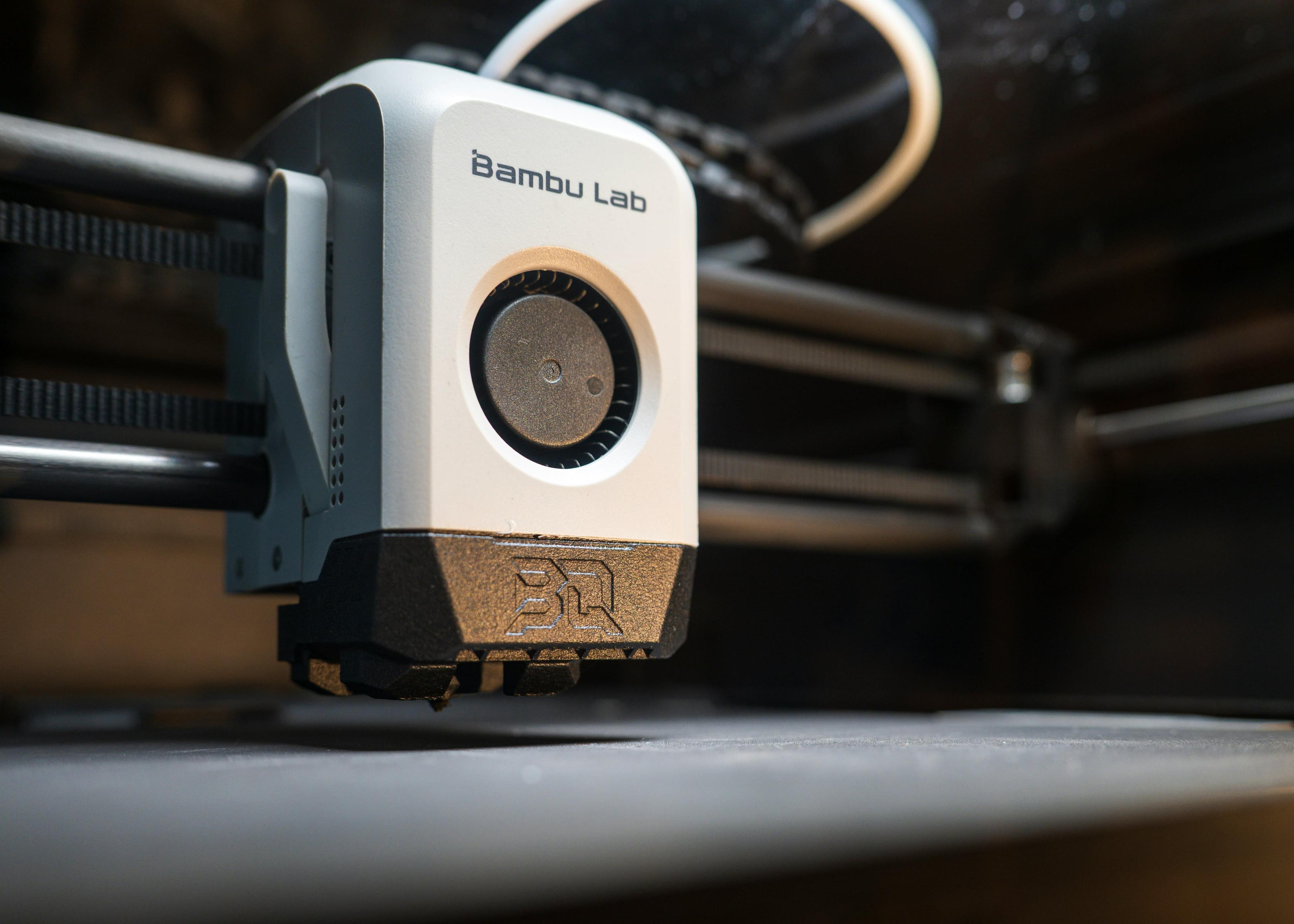 A Bambu Lab printer head, poised over a printing mad, against a dark background, with mounting rails visible.