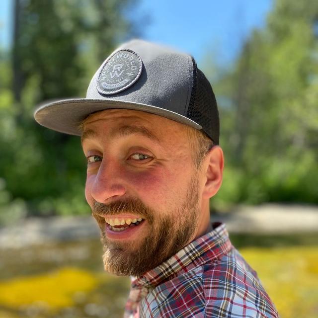 A smiling man with a beard wearing a gray and black baseball cap and a plaid shirt, standing outdoors on a sunny day with a blurred forest background.